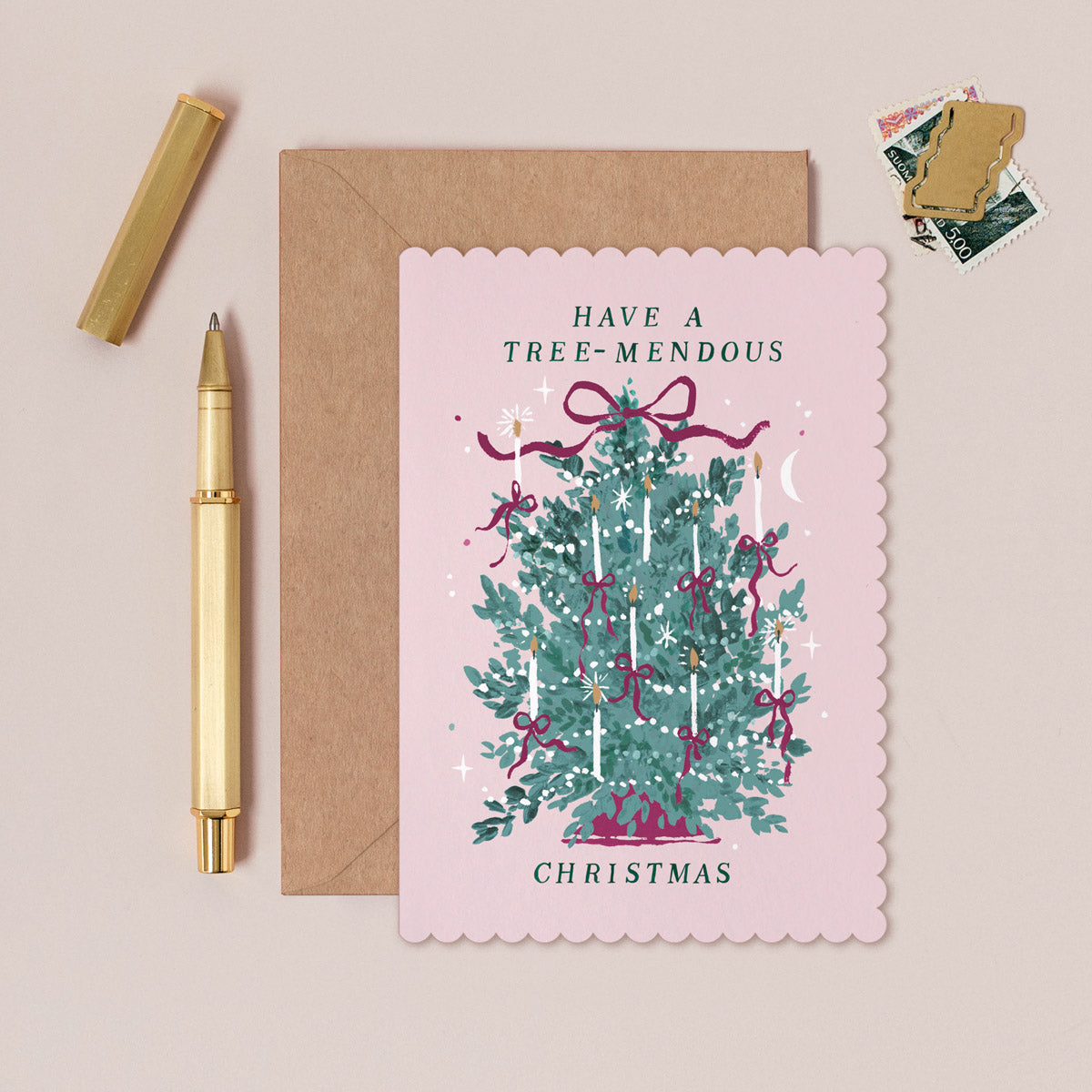 Christmas card with tree illustration and 'Have a Tree-mendous Christmas' text, next to a pen and envelope on a beige background.