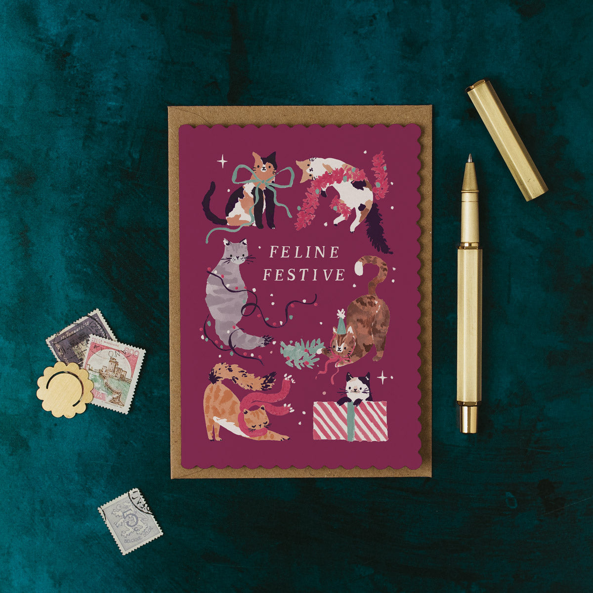 Feline Festive Christmas card illustrated with funny cats on a teal background with a pen and decorative items.