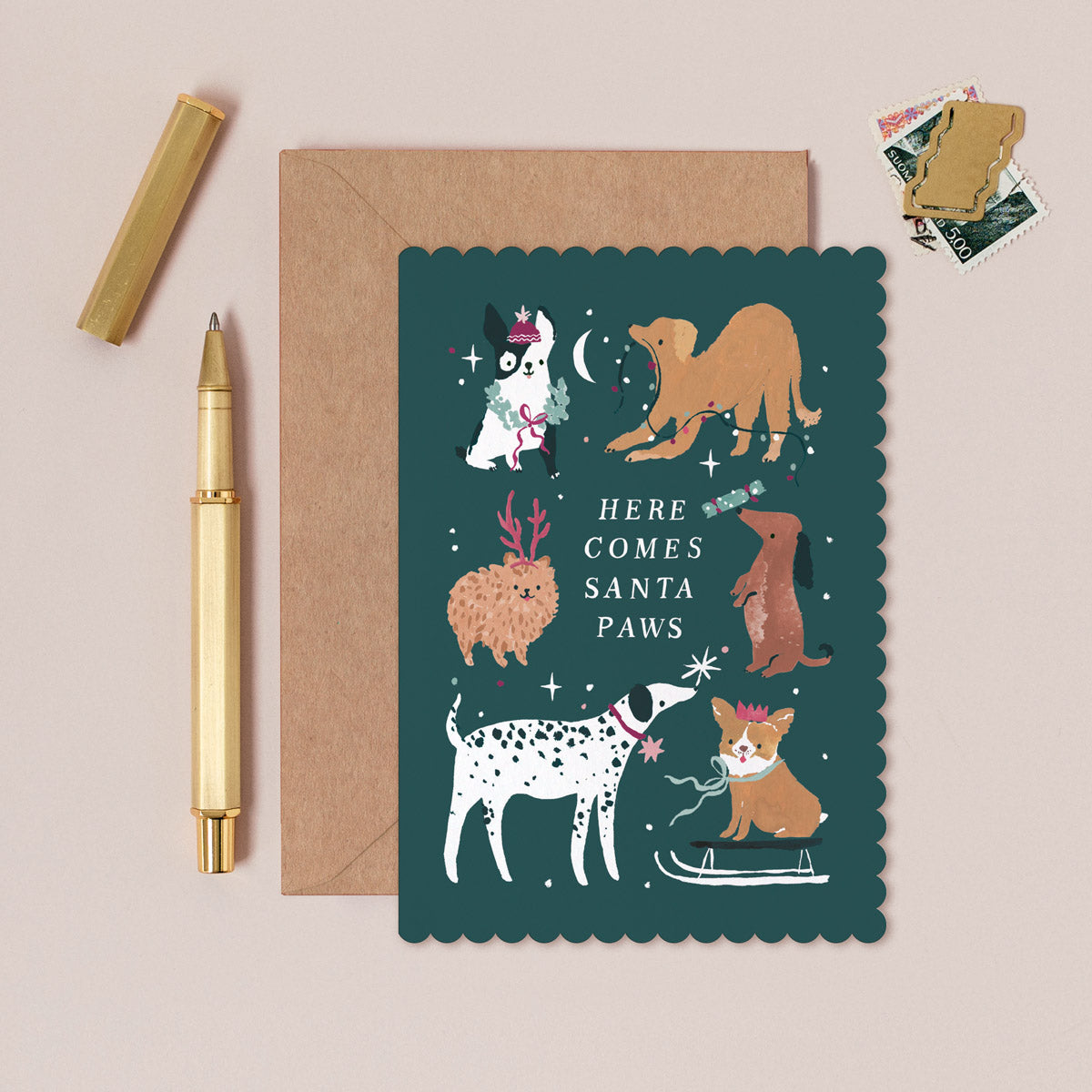 Christmas card with dog illustrations and 'Here Comes Santa Paws' text, next to a pen and envelope on a beige background.