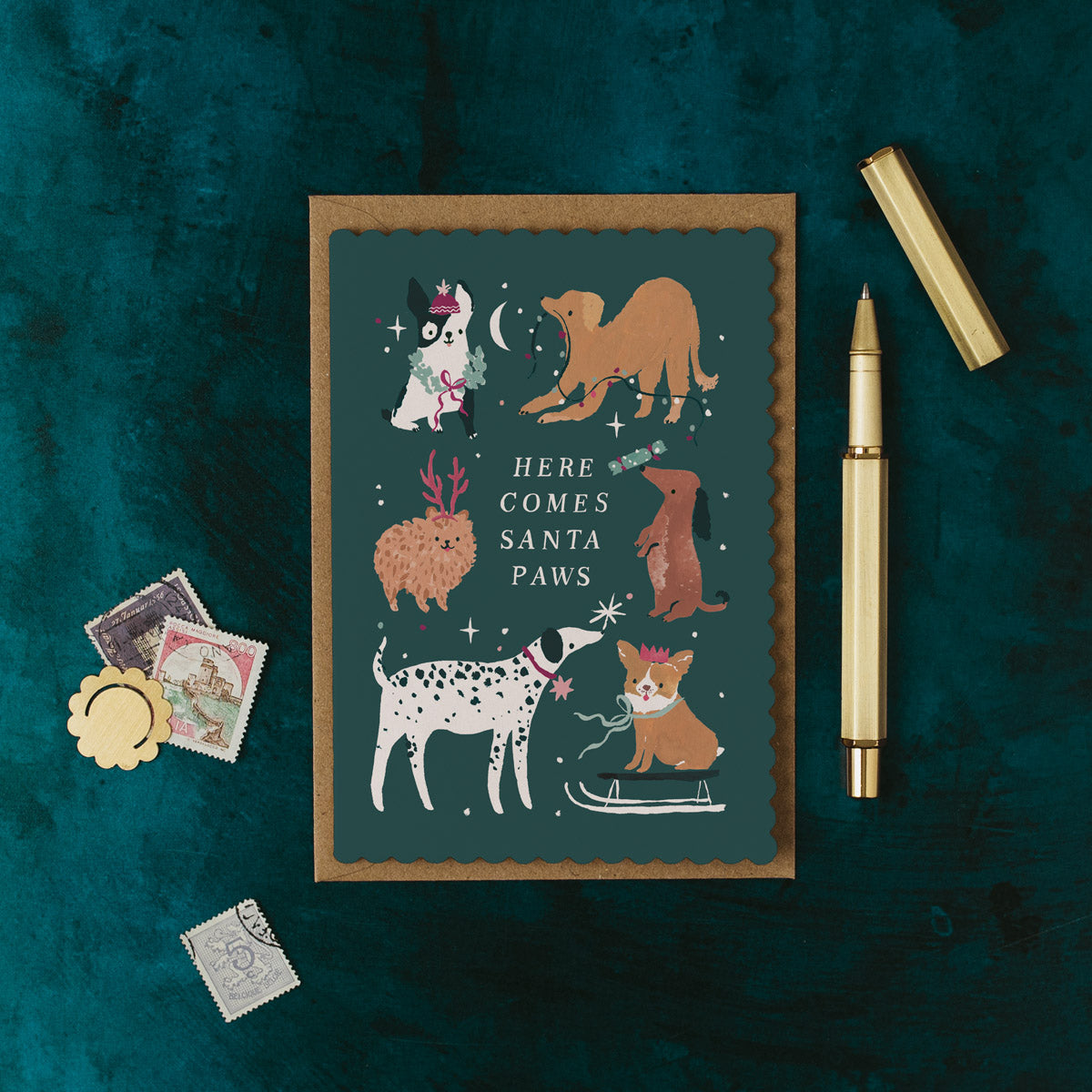 Christmas card with illustrations of dogs and text 'Here Comes Santa Paws' on a dark green background, with a pen and decorative items around.