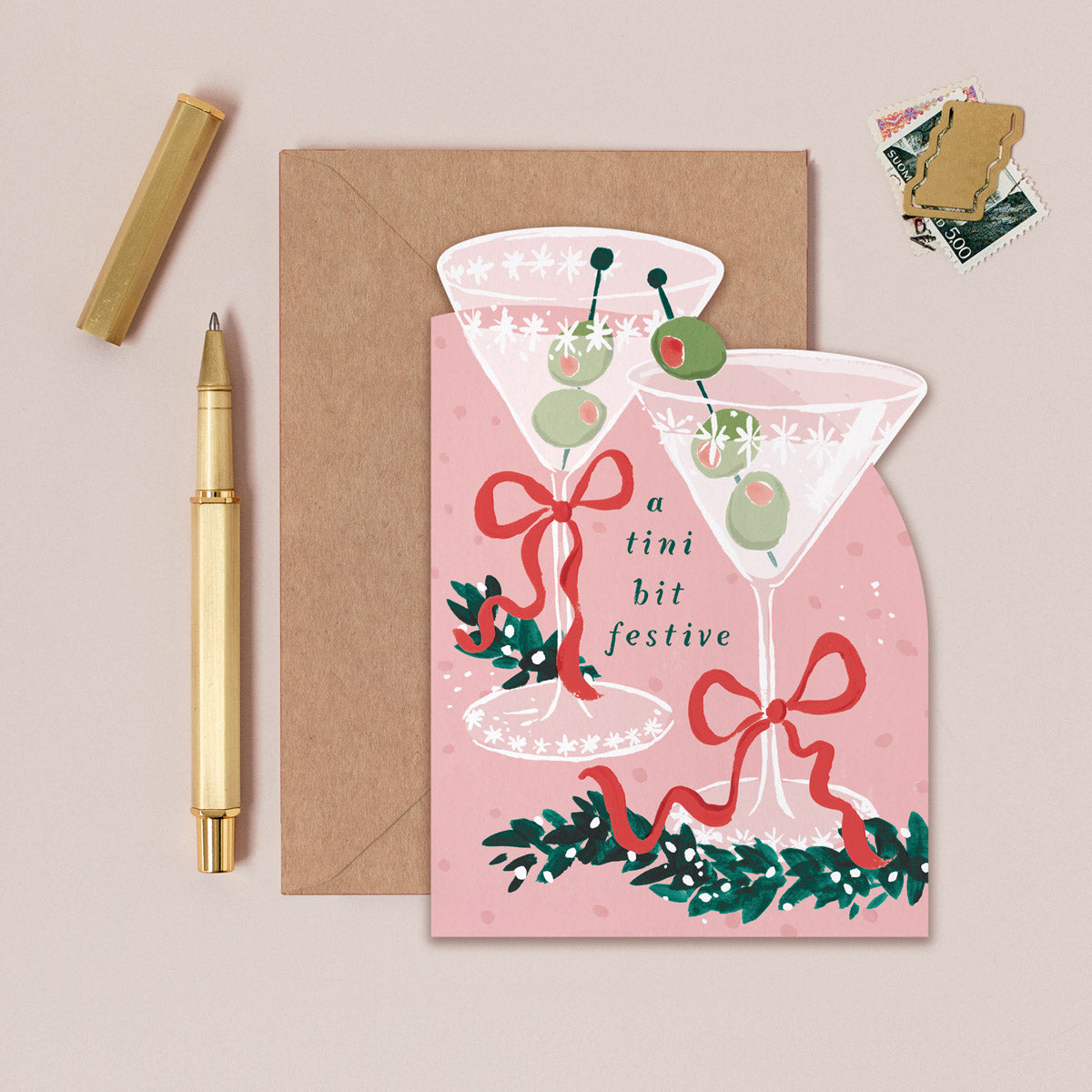 Christmas card with festive cocktail design, envelope, pen, and stickers on a beige background