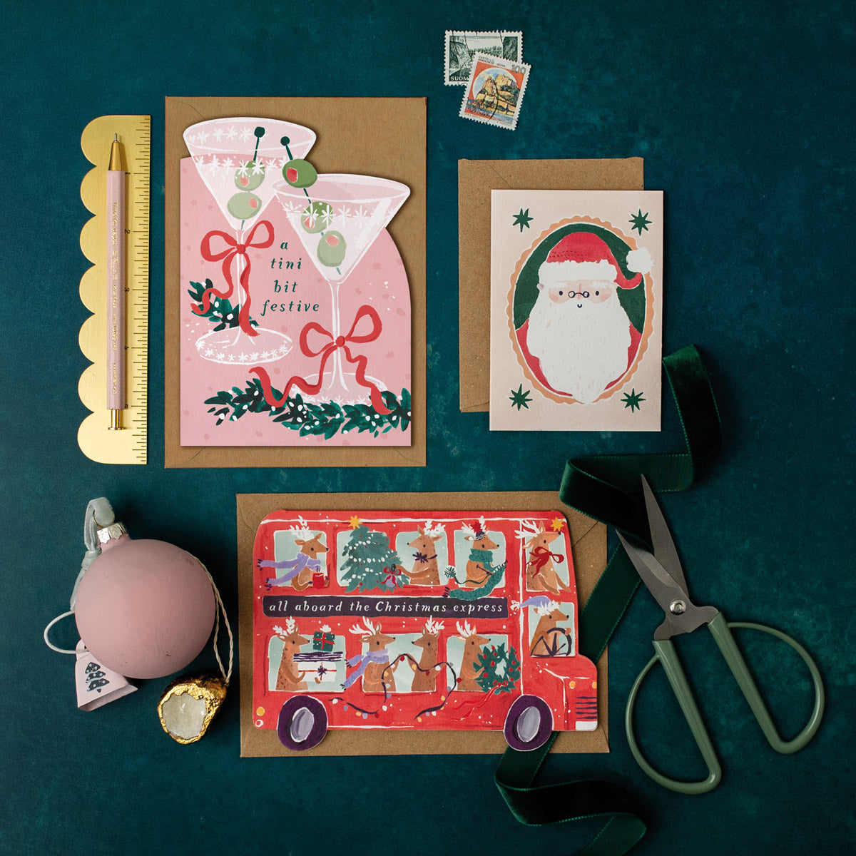 Three Christmas cards on a dark green surface with stationery items including a martini holiday card