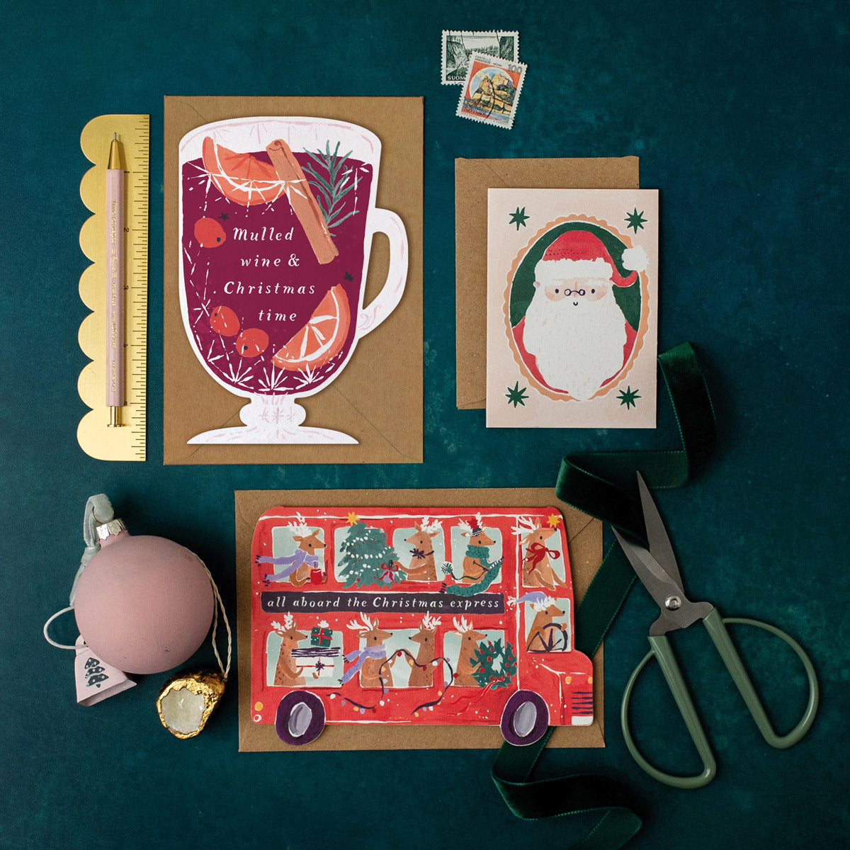 Three Christmas cards on a dark green surface with stationery items including a mulled wine shape holiday card.