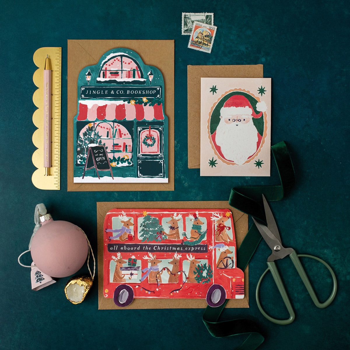 Three Christmas cards on a dark green surface with stationery items including a bookish bookshop Christmas card.
