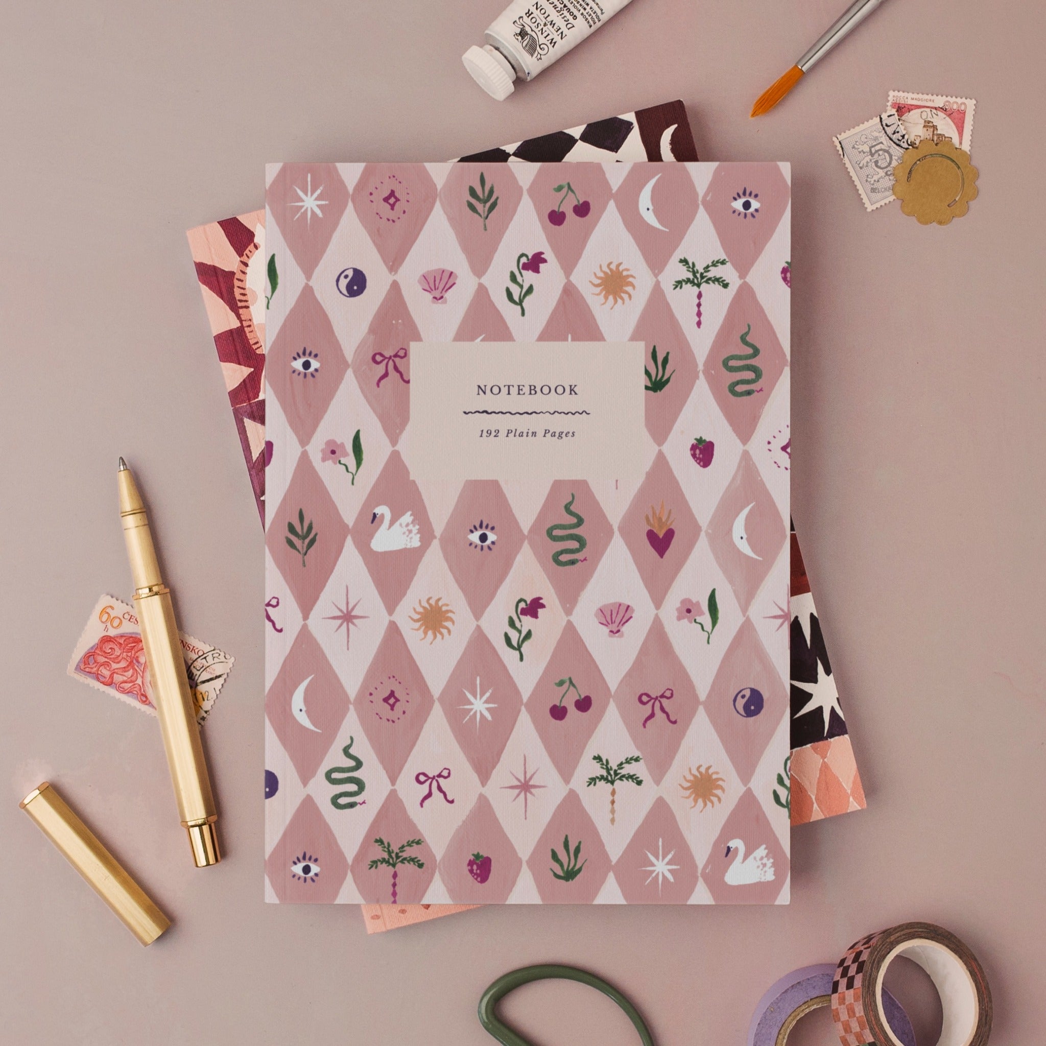 Blush Harlequin notebook, A5 plain page journal styled in a flat lay with a gold pen, paints and vintage stamps