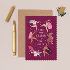 Christmas card with illustrations of women dancing and text 'TITS THE SEASON TO BE JOLLY' on a pink background, with a gold pen and envelope.