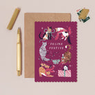 Feline Cat Christmas card with cats in festive attire on a pink background, accompanied by a gold pen and brown envelope.