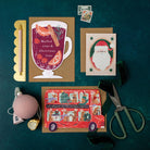 Three Christmas cards on a dark green surface with stationery items including a mulled wine shape holiday card.
