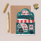 Bookish Christmas card with a festive bookshop illustration, envelope, pen, and stickers on a beige background