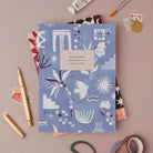 Medina Blue notebook, A5 plain page journal styled in a flat lay with a gold pen, paints and vintage stamps