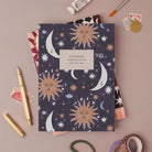 Sun and Moon notebook, A5 plain page journal styled in a flat lay with a gold pen, paints and vintage stamps
